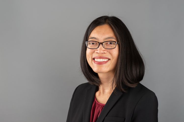 A professional headshot of a woman wearing a black blazer and red shirt with black glasses. 