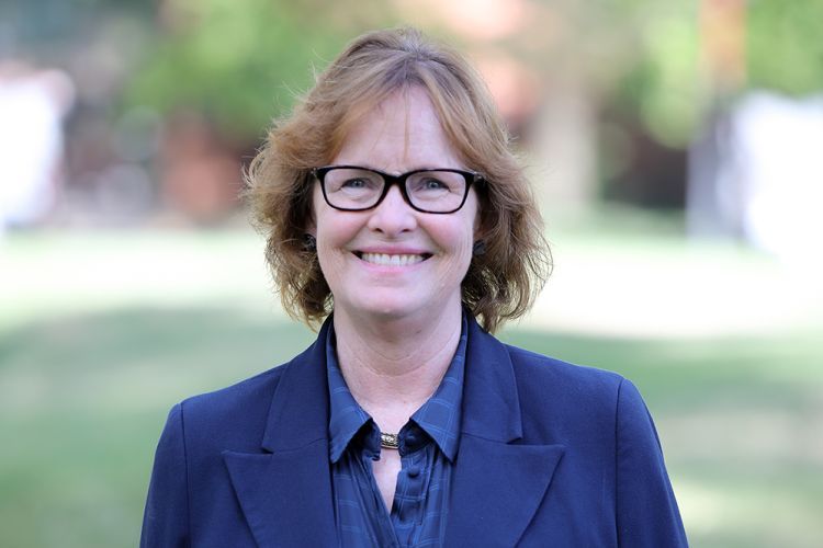 A woman with brown curly hair wearing a blue blazer stands outside for a professional photo. 