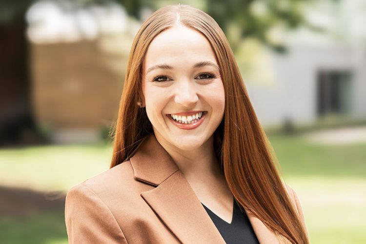 A women with long ginger hair wearing a brown suit jacket and black top. 