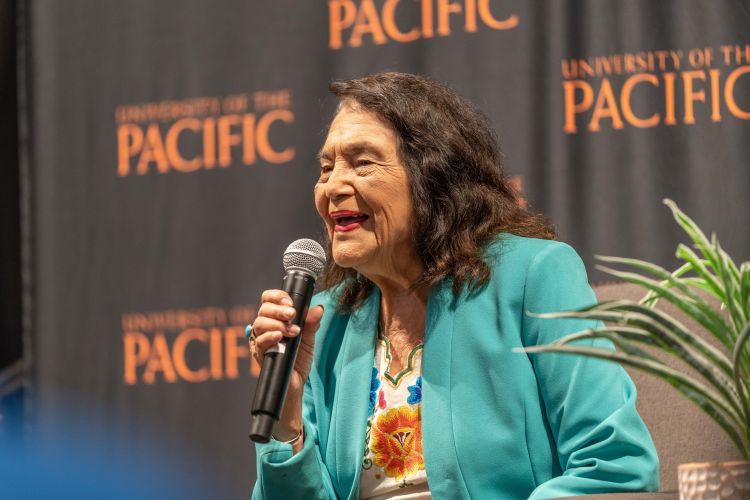 Dolores Huerta smiling with a microphone in hand and a University of the Pacific backdrop in the foreground. 