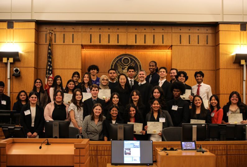 A group of high school students pose for a photo in a courtroom with a few judges