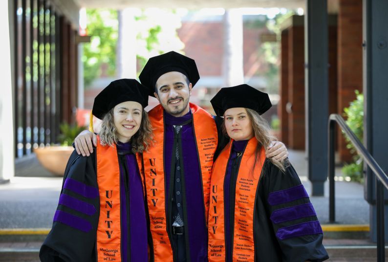 Four graduates wear purple and black robes with graduation caps on and orange sashes that say "UNITY McGeorge School of Law"