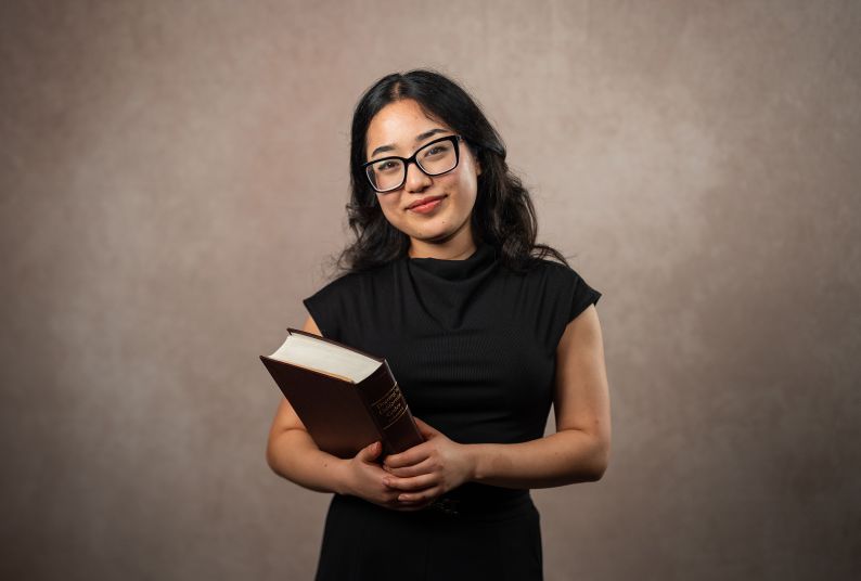 A student holding a textbook in front of a neutral background 