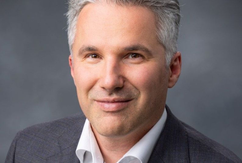 A professional portrait of a man wearing a grey suit against a dark background. 