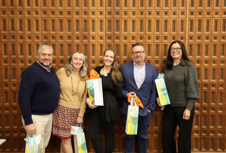 Five people holding gift bags stand in front of a wooden wall smiling for a picture. 