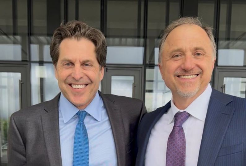 Two men in ties, collared shirts and blazers stand and smile for a photo. 