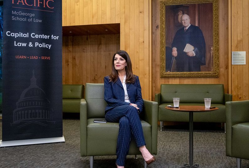 A woman in a dark blue stripped suit sits in a green chair in front of a sign that says "University of the Pacific McGeorge School of Law Capital Center for Law and Policy."
