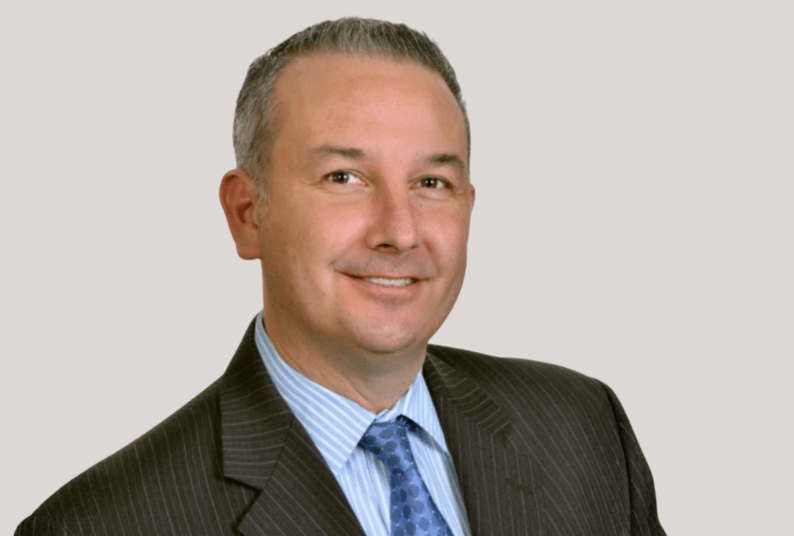 A headshot portrait of a man wearing a grey suit jacket a blue tie and a light blue under shirt. He has short gray hair and brown eyes.  