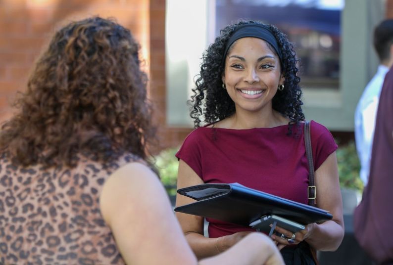 Two women in business attire conversing. One is smiling and holding a binder