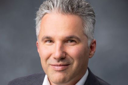 A professional portrait of a man wearing a grey suit against a dark background. 