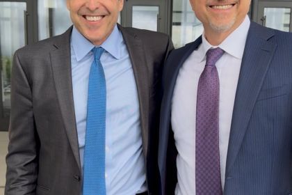Two men in ties, collared shirts and blazers stand and smile for a photo. 