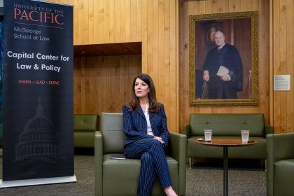 A woman in a dark blue stripped suit sits in a green chair in front of a sign that says "University of the Pacific McGeorge School of Law Capital Center for Law and Policy."