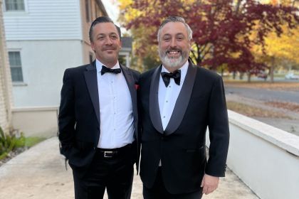 Two brothers stand in an outdoor setting wearing nice suits smiling together.