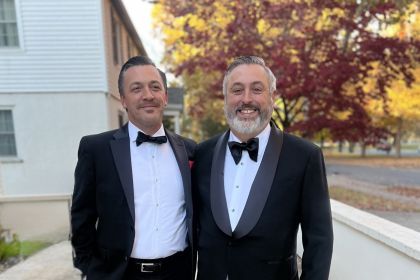 Two brothers stand in an outdoor setting wearing nice suits smiling together.