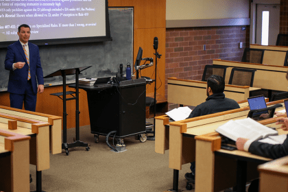 A professor wearing a blue suit stands in front of a lecture hall of students teaching a class.