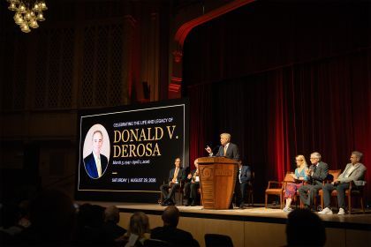 several people sit on a stage next to a large screen that reads "Celebrating the Life and Legacy of Donald V. DeRosa"