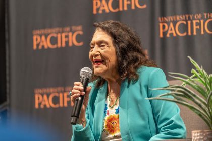 Dolores Huerta smiling with a microphone in hand and a University of the Pacific backdrop in the foreground.