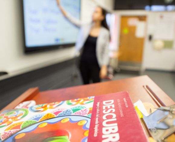 Spanish textbook in the foreground; someone teaching in the background