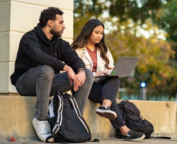 two students sitting outdoors and looking at a laptop screen