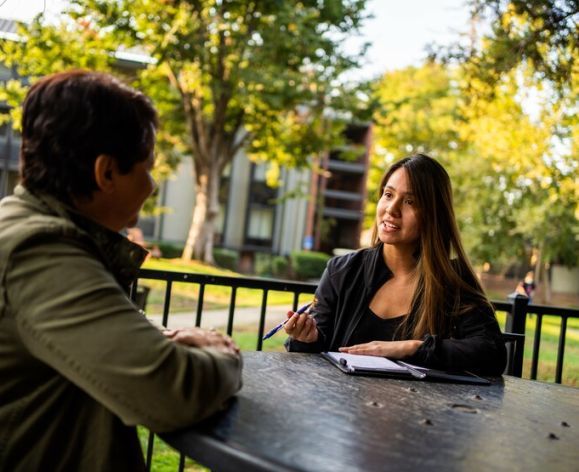 Photo shows two students talking at an outdoor table on the Sacramento campus.