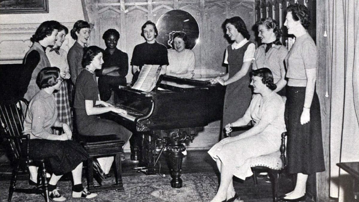 A group of women stand around a piano in South Hall.