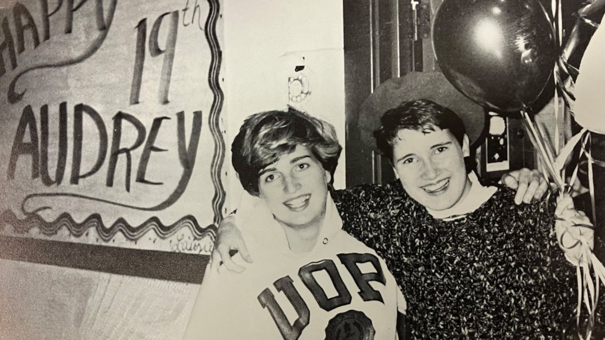 Two friends stand in front of a "Happy Birthday" sign with balloons.